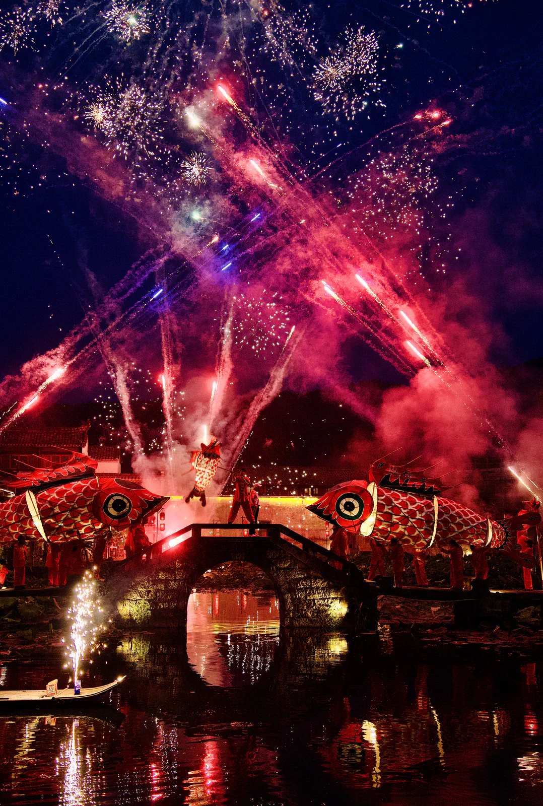 Fireworks bursting above a stone bridge and glowing fish lanterns.