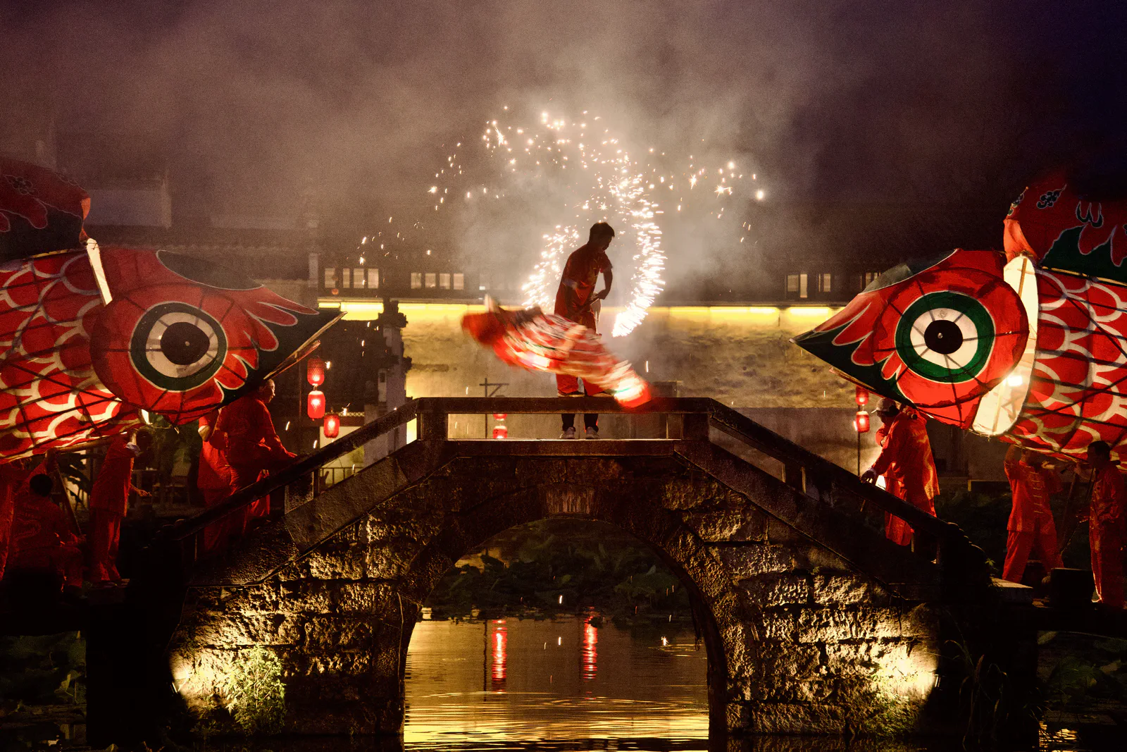 A performer surrounded by sparks on a stone bridge between fish lanterns.