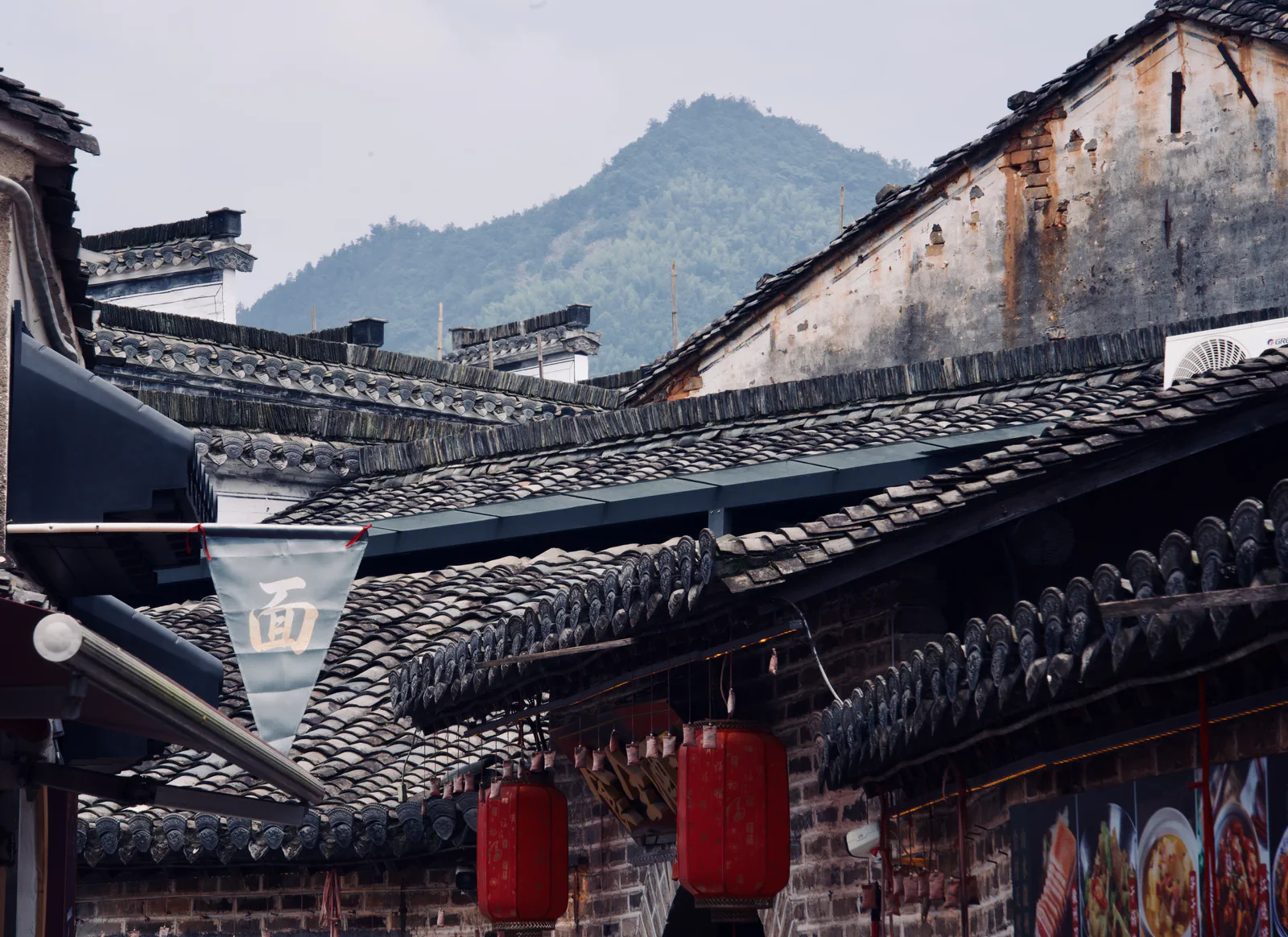 Old tiled rooftops and red lanterns with a green mountain behind them.