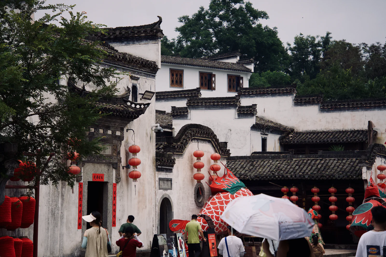 Visitors carrying umbrellas outside a white-walled entrance decorated with red lanterns.