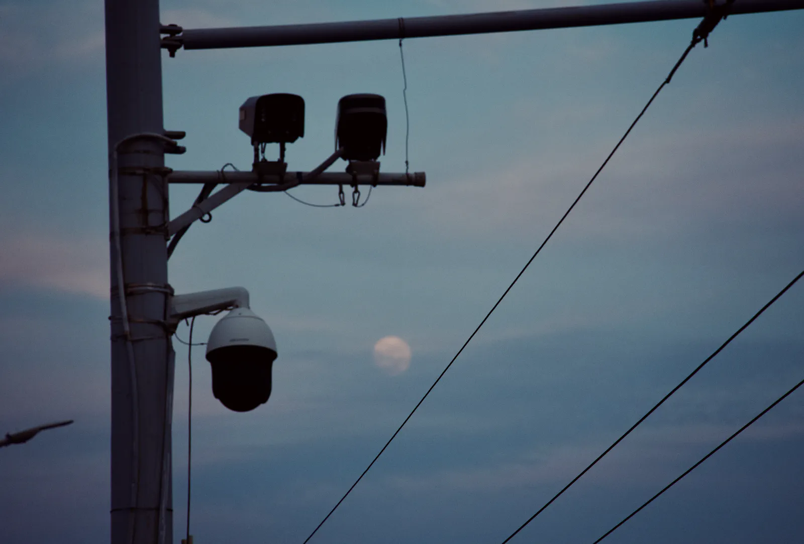 A pale moon seen through overhead wires beside a riverside pole.