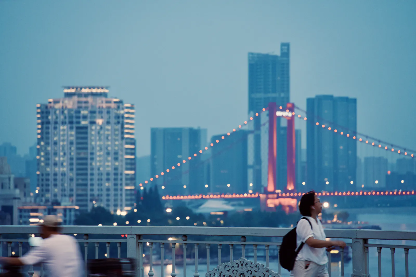 Pedestrians on the waterfront with a lit suspension bridge in the distance.