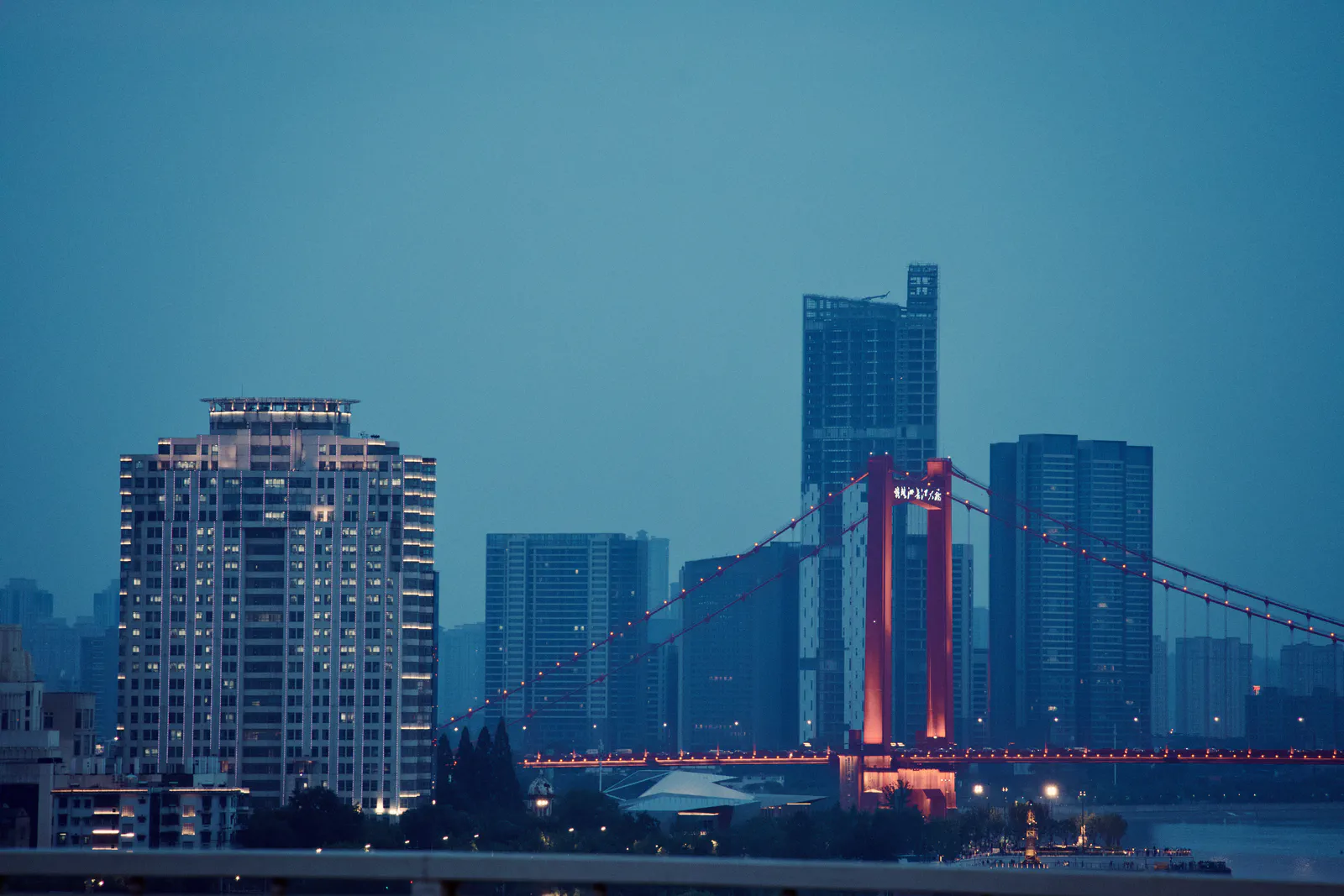 A suspension bridge and dense city buildings across the river at blue hour.