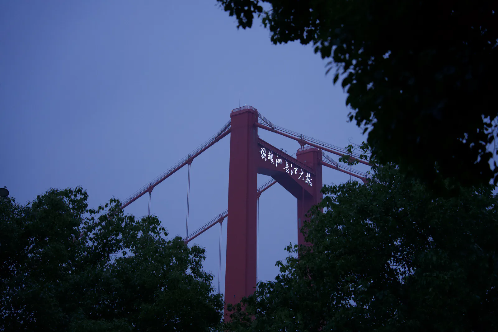A red suspension bridge tower framed by leafy trees at dusk.