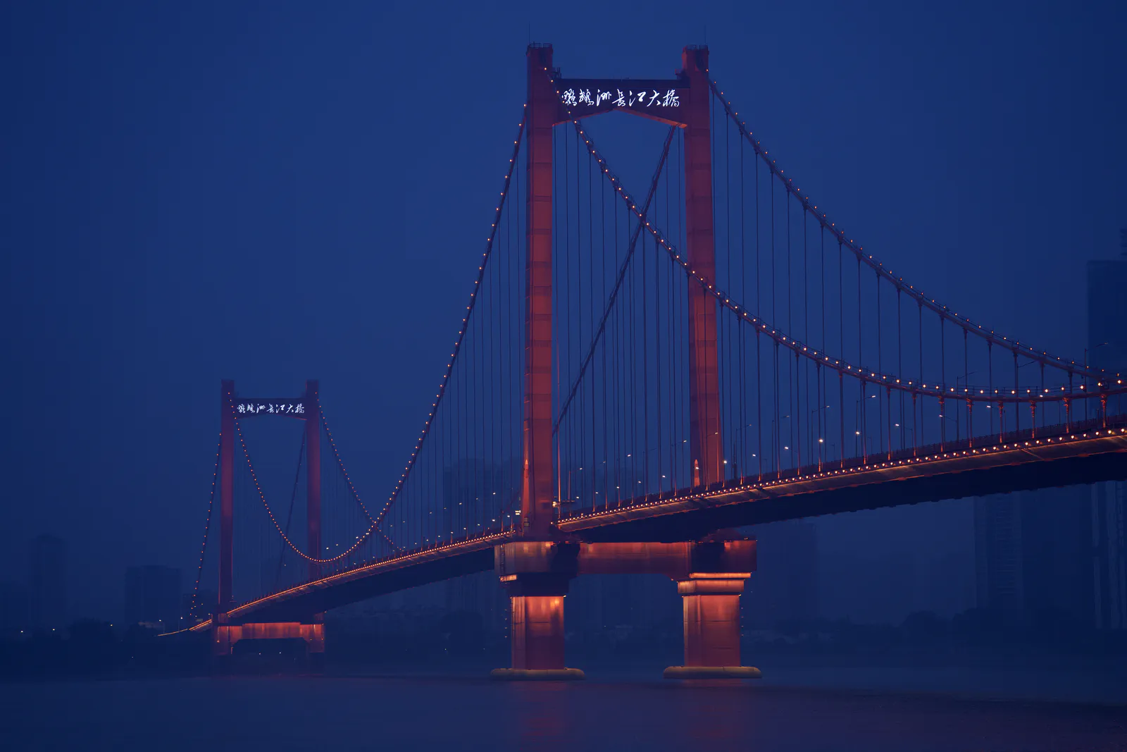 An illuminated red suspension bridge stretching across the river at blue hour.