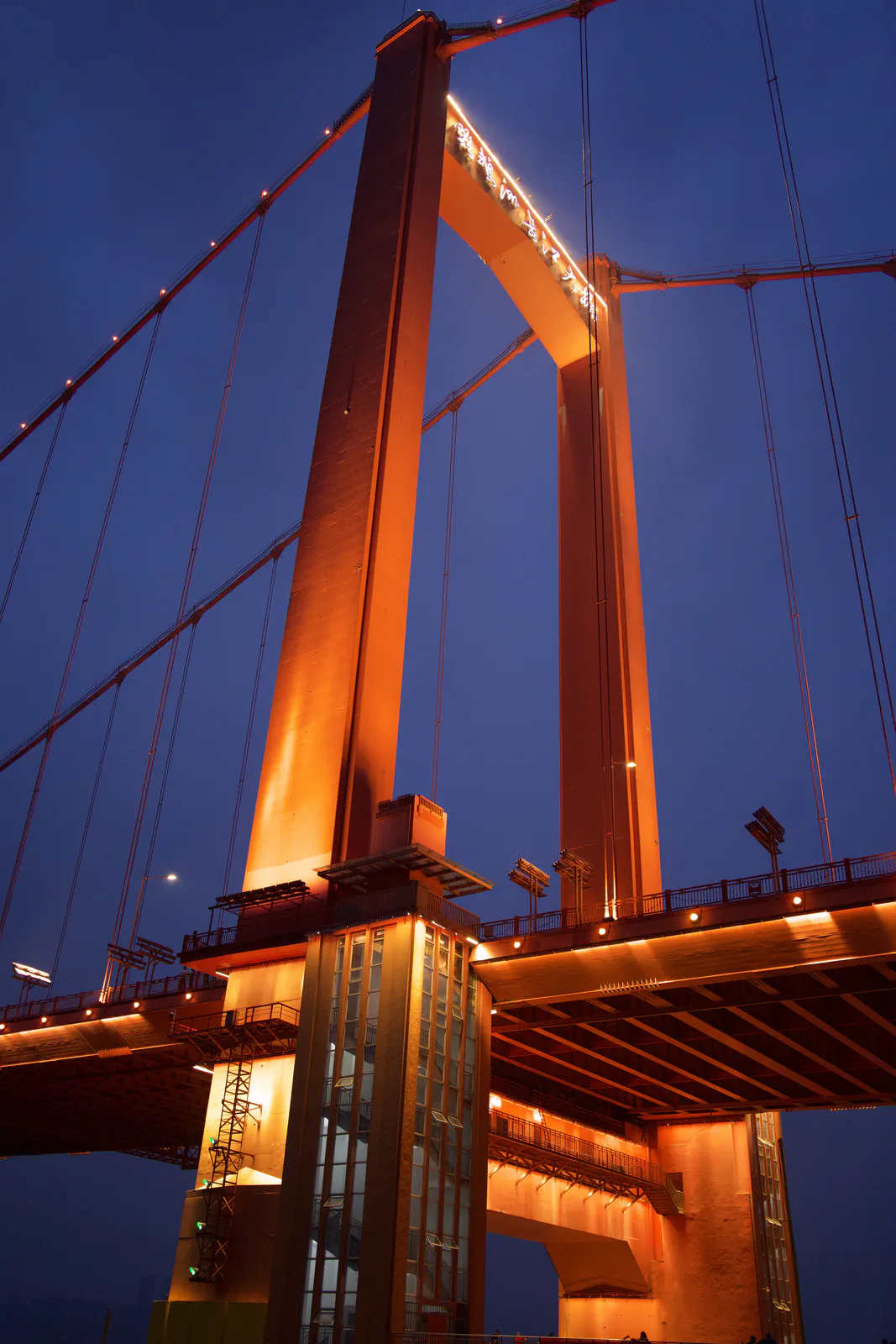 A close upward view of a lit bridge tower and suspension cables.