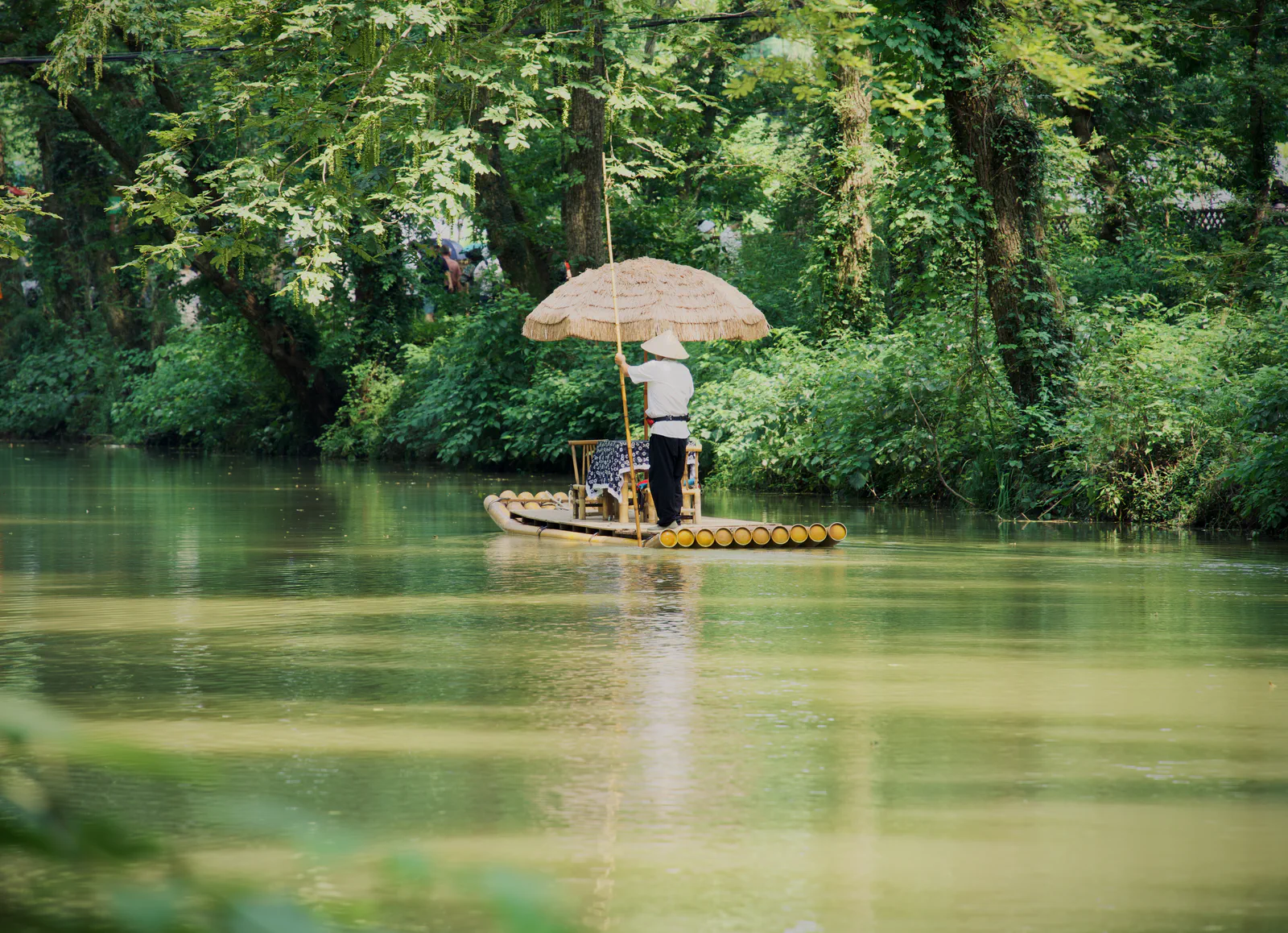 A person beneath a pale umbrella on a bamboo raft beside lush riverside trees.